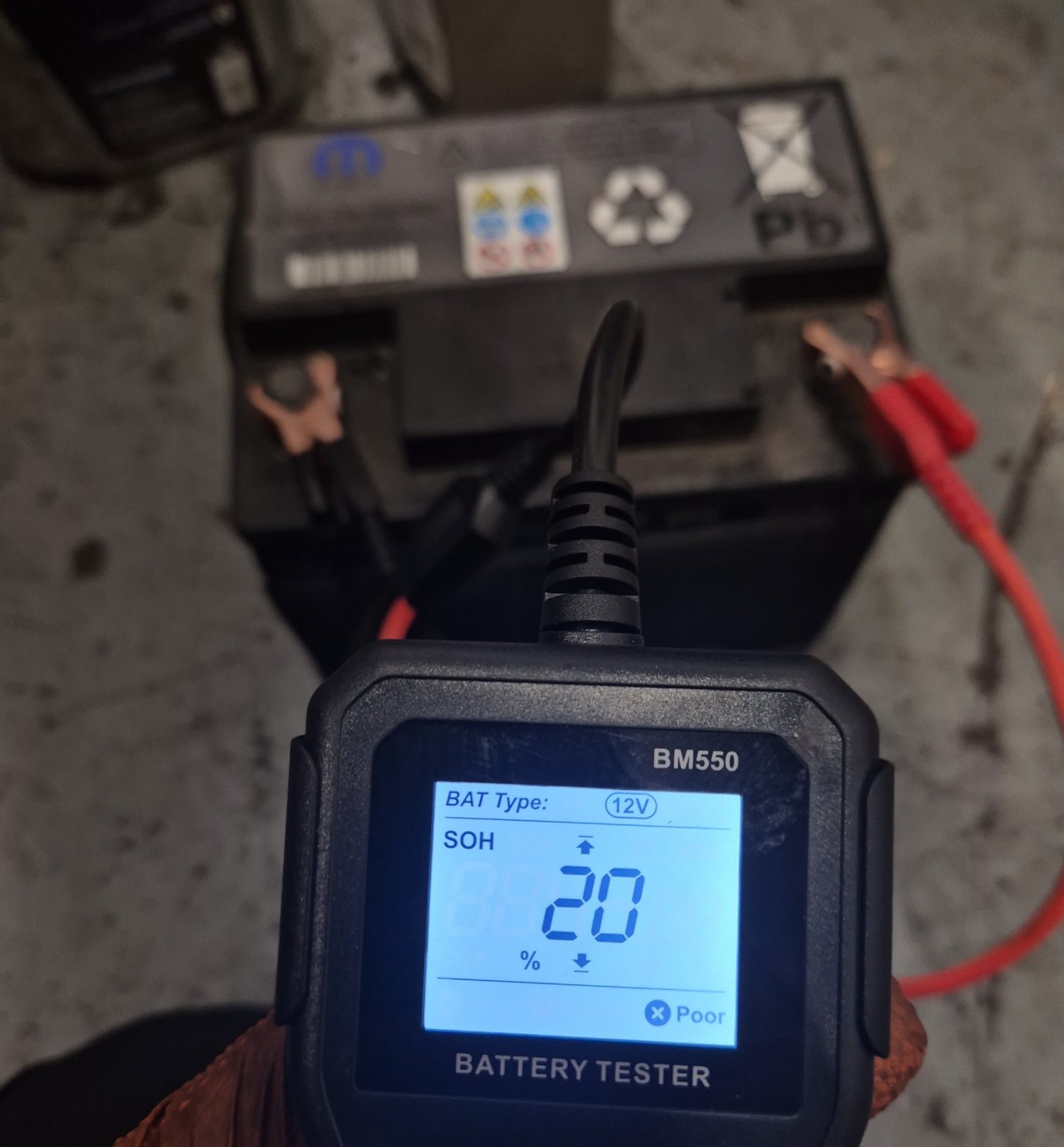 A BM550 battery tester clipped to a car battery, reading 20 percent state of health