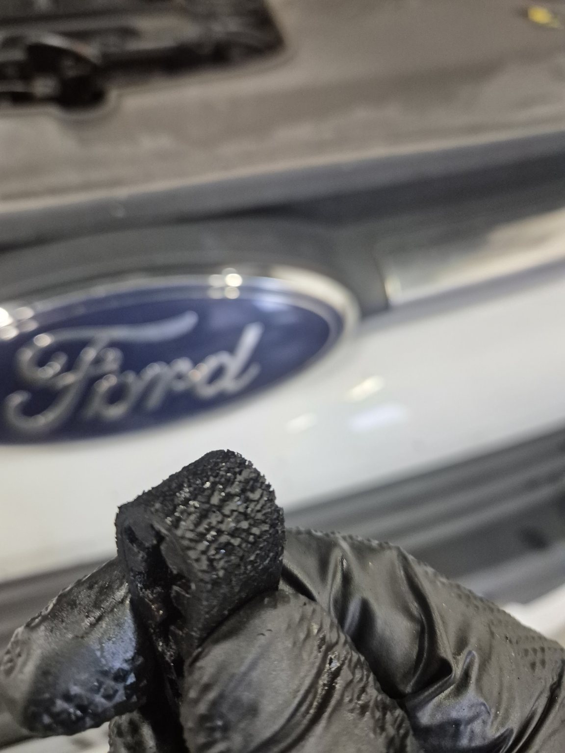 A gloved hand holding a worn, broken part up in front of a Ford badge